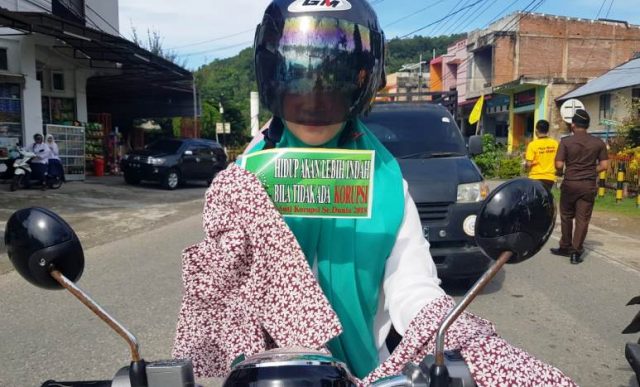 WhatsApp Image 2018-12-10 at 15.04.57 Seorang wanita berkenderaan memegang stiker anti korupsi, sementara tampak di pinggir jalan para pegawai Kejaksaan Aceh Selatan membagikan kaos dan stiker Anti Korupsi kepada pengguna jalan lainnya, di Simpang Kedai Haru, Tapaktuan, Aceh Selatan, Senin (10/12/2018). (Foto/Faisal).